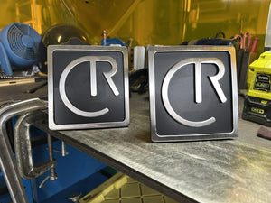 Two metal signs with 'CR' logo on a workshop counter.